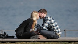 Ein junges Paar auf einer Mauer sitzt sich im Schneidersitz gegenüber und küsst sich.