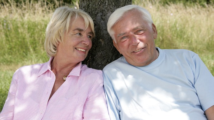 Seniorenpaar lehnt sich an einen Baum. Sie schaut ihn lächelnd von der Seite an.
