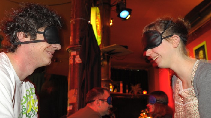 Mann und Frau sitzen sich mit verbundenen Augen in einem Club gegenüber und unterhalten sich | Bildquelle: dpa Picture-Alliance / Soeren St. Mann und Frau sitzen sich mit verbundenen Augen in einem Club gegenüber und unterhalten sich