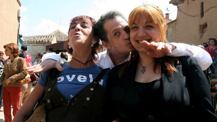 Mann in spanischem Dorf umarmt zwei Frauen von hinten und küsst eine auf die Wange.