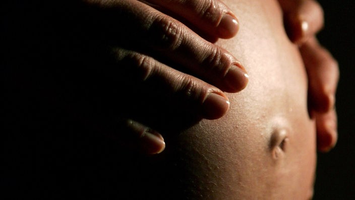 Eine hochschwangere Frau fasst sich mit beiden Händen an ihren Bauch.