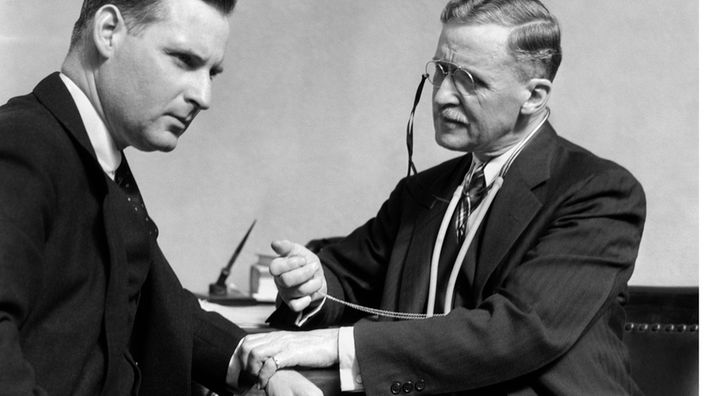 Schwarzweiß-Foto: Ein Arzt fühlt einem Patienten im Anzug den Puls.