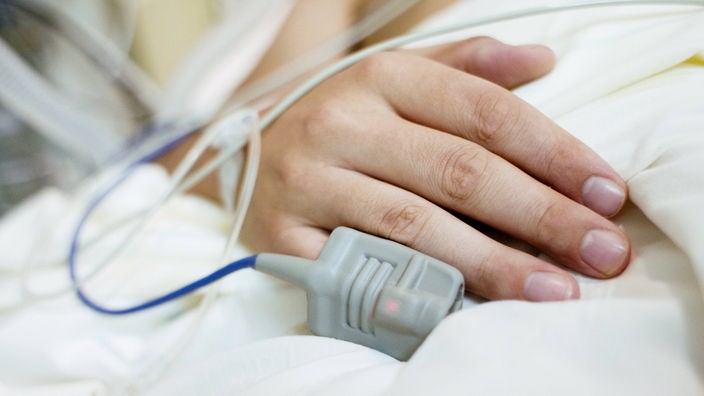 Die Hand eines Patienten liegt auf einem Bett, am Finger befindet sich ein Sauerstoffmessgerät