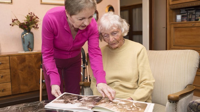 Eine Frau zeigt einer Seniorin ein Buch mit Fotos. Die Szenespielt sich in einer häuslichen Umgebung ab.