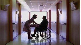 Pflegerin mit Mann im Rollstuhl auf dunklem Flur.