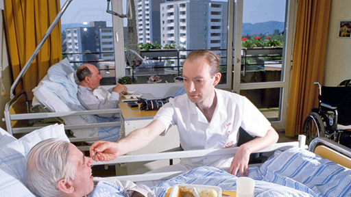 Pfleger füttert einen Pflegebedürftigen in einem Mehrbettzimmer.