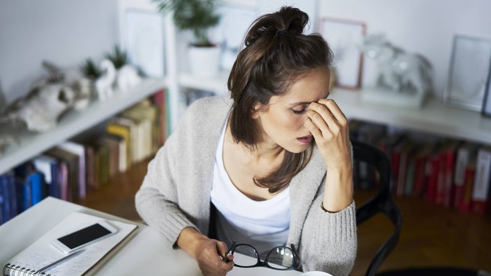 Eine Frau am Schreibtisch reibt sich müde die Augen, vor ihr eine Tasse kaffee und Tabletten.