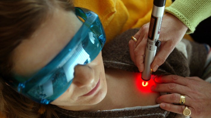 Eine Patientin, die eine spezielle Brille trägt, um ihre Augen vor der Laserstrahlung zu schützen, wird an einem Akupunkturpunkt im Bereich der Brust mit einem Laserstift behandelt.