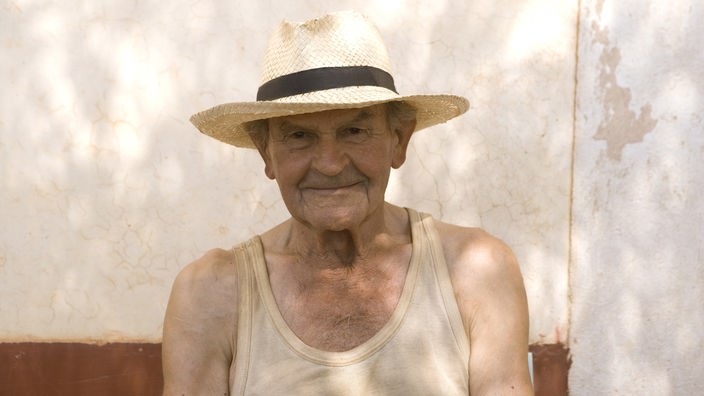 Das Bild zeigt einen älteren Mann, der vor einer Hausmauer sitzt. Er trägt einen Strohhut und ist nur mit einem Unterhemd bekleidet.