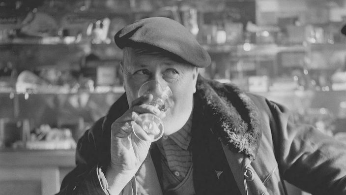 Das Bild zeigt einen älteren Mann im Bistro. Er trägt eine Baskenmütze und trinkt einen Drink.