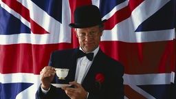 Das Bild zeigt einen Mann vor der Flagge Großbritanniens. Er trägt einen eleganten, schwarzen Anzug und einen Bowler, einen steifen Herrenhut aus schwarzem Filz mit halbkugelförmigem Kopf. In der Hand hält er eine Tasse.