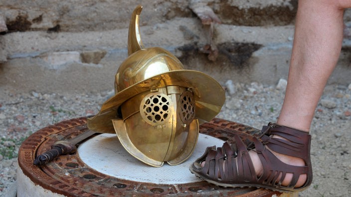 Römische Militärsandale mit Lederriemen neben einem goldenen Gladiatorenhelm.