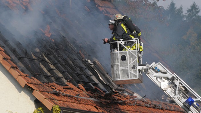Feuerwehrleute auf einer Leiter vor einem rußgeschwärzten Dach.
