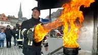 Auf einem Platz im Freien steht ein Feuerwehrmann mit einem Topfdeckel in der Hand. Aus dem Topf mit Fett schiessen riesige Flammen.