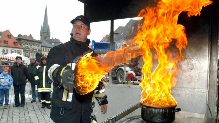Auf einem Platz im Freien steht ein Feuerwehrmann mit einem Topfdeckel in der Hand. Aus dem Topf mit Fett schiessen riesige Flammen.