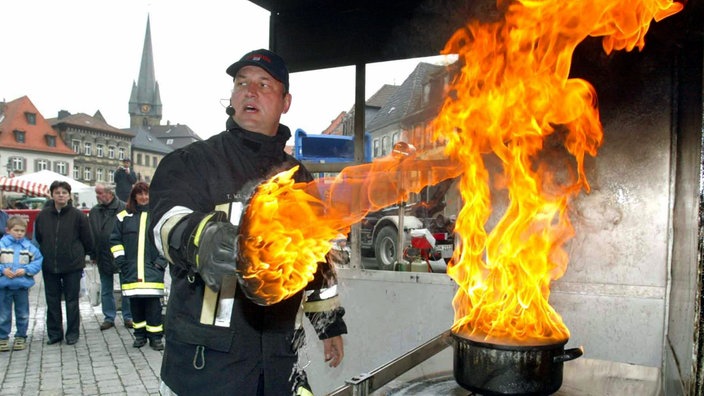 Auf einem Platz im Freien steht ein Feuerwehrmann mit einem Topfdeckel in der Hand. Aus dem Topf mit Fett schiessen riesige Flammen. | Bildquelle: dpa Auf einem Platz im Freien steht ein Feuerwehrmann mit einem Topfdeckel in der Hand. Aus dem Topf mit Fett schiessen riesige Flammen.