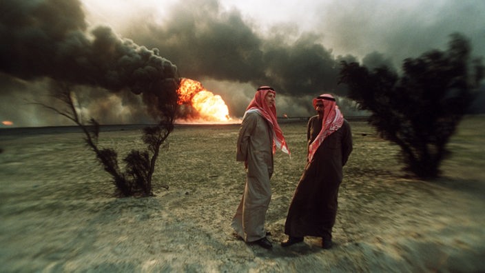 In einer sandigen, flachen Wüstenlandschaft stehen zwei traditionell gekleidete Kuwaitis vor einer brennenden Ölquelle.