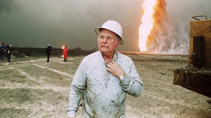 Red Adair mit weißem Helm in der irakischen Wüste. Im Hintergrund eine brennende Ölquelle. | Bildquelle: dpa/AFP Red Adair mit weißem Helm in der irakischen Wüste. Im Hintergrund eine brennende Ölquelle.