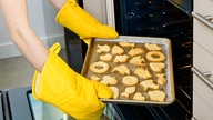 Zwei Hände in gelben Topflappen ziehen ein Blech mit Weihnachtsplätzchen aus einem Herd.
