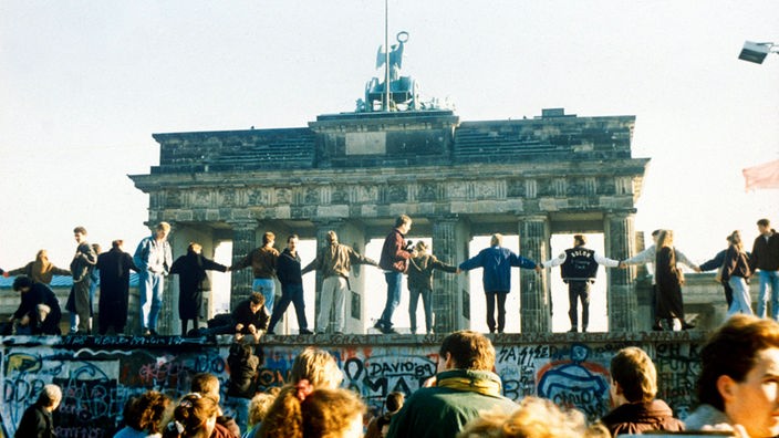 Viele Menschen stehen auf der Berliner Mauer und halten sich an den Händen. Im Hintergrund: das Brandenburger Tor.