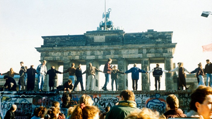 Viele Menschen stehen auf der Berliner Mauer und halten sich an den Händen. Im Hintergrund: das Brandenburger Tor.
