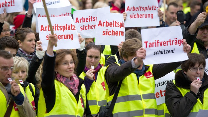 Gewerkschafterinnen und Gewerkschafter in gelben Westen halten Schilder hoch, auf denen steht: "Arbeitsplatzsicherung" und "Tarifverträge schützen!"