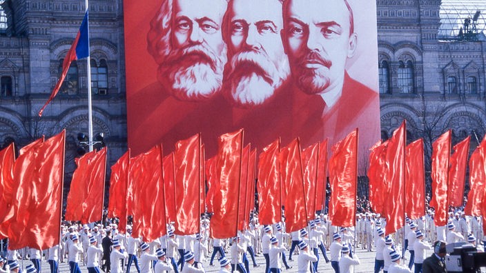 Hinter einer marschierenden Menschenmenge in Moskau mit roten Fahnen hängt ein  riesiges Bild mit Karl Marx, Friedrich Engels und Wladimir Iljitsch Lenin