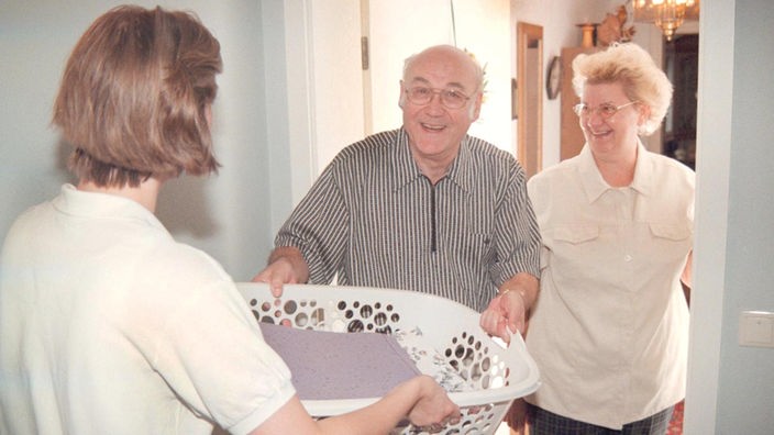 In einer offenen Wohnungstür steht lächelnd ein älteres Paar. Der Mann nimmt einen weißen Korb mit Wäsche von einer blonden Frau im Flur entgegen.