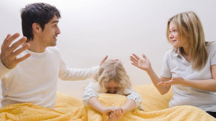 Ehepaar streitet im Bett, Kind sitzt hilflos dazwischen.