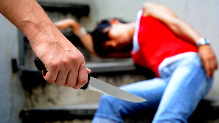 Mann hält Messer in der Hand, im Hintergrund liegt eine Frau auf Treppen.