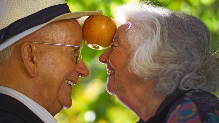 Ein Seniorenpaar balanciert zwischen den Köpfen einen Apfel.