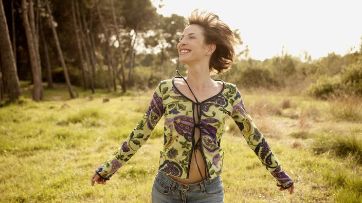 Lachende Frau läuft springend über eine Wiese im Sonnenschein.
