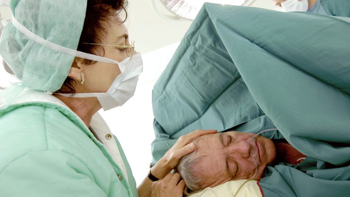 Eine Frau befindet sich in einem OP-Saal und legt ihre Hand auf den Kopf eines Patienten