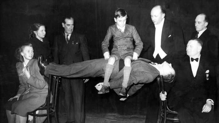 Schwarzweiß-Bild: Eine Person liegt auf zwei Hockern, den Rücken in der Luft durchgestreckt. Ein Junge sitzt auf dem Bauch des Mannes.