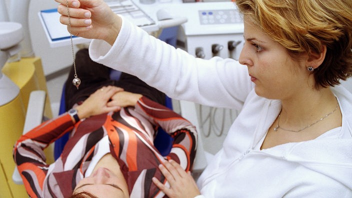 Eine Frau in einer Zahnarztpraxis hält ein Pendel über ihre liegende Patientin