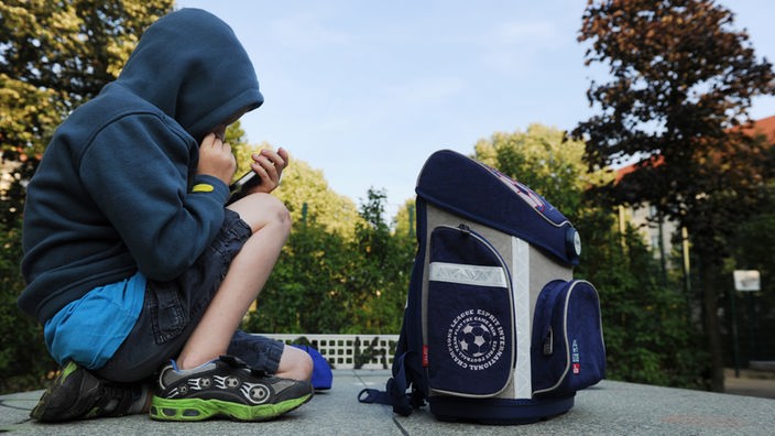 Ein Junge sitzt mit seinem Schulranzen auf einer Tischtennisplatte auf einem Spielplatz und spielt auf einem Smartphone