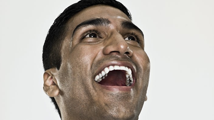 Ein junger, indischer Mann lacht lauthalt. Er trägt ein weißes Hemd und ist vor weißem Hintergrund abgelichtet.