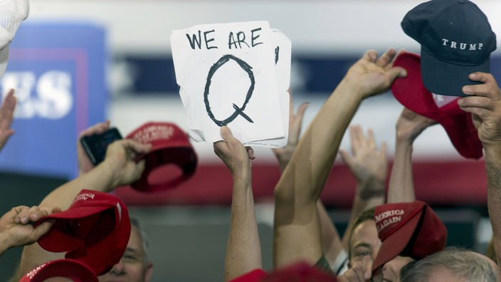 Menschen bei einer Demonstration; einer hält ein selbstgeschriebenes Blatt Papier mit der Aufschrift 'We are Q" in die Luft.