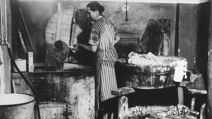 Das Schwarzweißbild zeigt eine Frau, die Wäsche mit einem Holzstab in einen Zuber befördert.