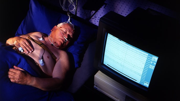 Ein Mann liegt schlafend in einem Bett. Er trägt eine Sauerstoffmaske auf der Nase und mehrere Kabel im Gesicht.