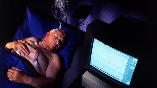 Ein Mann liegt im Schlaflabor mit Elektroden am Kopf und einem Computerbildschirm neben dem Bett.