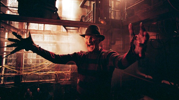 Freddy Krueger, ein Mann mit vernarbtem Gesicht, Hut und einem Messerhandschuh, steht schreiend in einer Fabrikhalle.