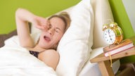 Eine junge Frau räkelt sich kurz nach dem Aufwachen in ihrem Bett. Auf einem Nachttisch steht ein Wecker