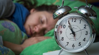 Im Vordergrund zeigt ein Wecker kurz vor 7 Uhr an, während im Hintergrund eine Frau im Bett liegt und schläft