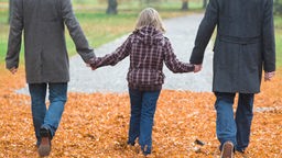 Ein schwules Pärchen geht mit einer Tochter in der Mitte in einem Park spazieren. Der Rücken ist jeweils der Kameraa zugewandt