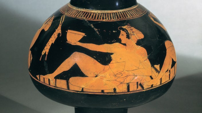 Malerei auf antiker Vase: Frau kauert auf Teppich und trinkt aus Gefäß.