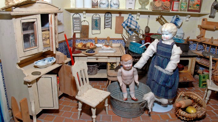 Puppenküche aus Deutschland um 1920. Eingerichtet mit Schrank, Herd und Tisch und verschiedenen Töpfen und Geschirr. Eine Puppe, einem Jungen nachempfunden, sitzt in einer Zinkwanne. Daneben steht eine weitere Puppe, die die Mutter darstellt.