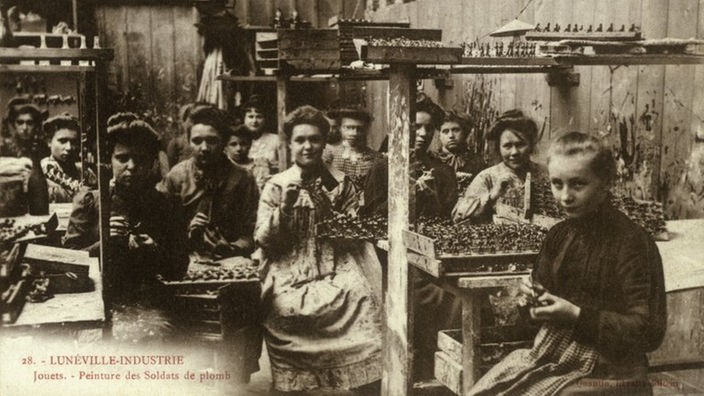 Eine Schwarzweiß-Fotografie im Postkartenformat zeigt einen Raum, in dem von circa zwölf Frauen Zinnsoldaten hergestellt und bemalt werden. Das Foto stammt aus Frankreich und ist auf das Jahr 1900 datiert.