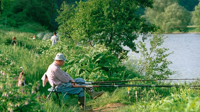 Angler sitzt neben mehreren Angelruten am Ufer