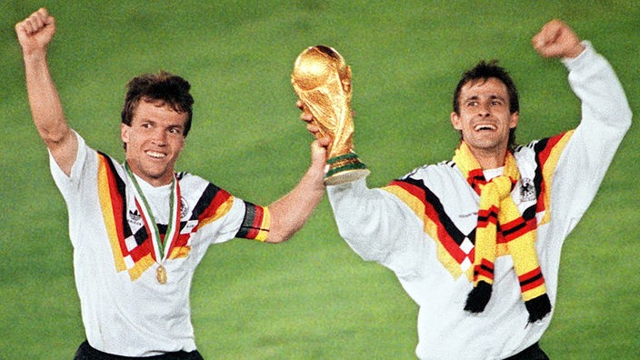 Lothar Matthäus und Pierre Littbarski laufen nebeneinander über das Spielfeld. Zwischen sich halten sie den WM-Pokal mit hoch erhobenen Händen.